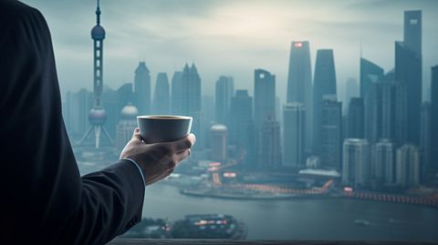 A closeup of hands holding a steaming cup of coffee with the Shanghai skyline in the background, symbolizing the company's presence in the People's Republic of China.